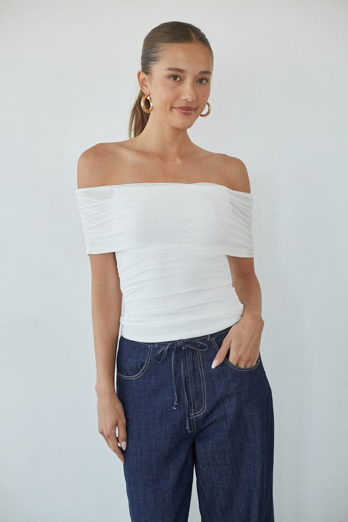Woman wearing a white off-shoulder top and blue jeans against a plain background