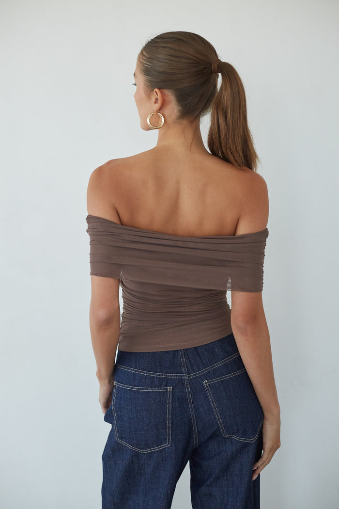 Woman wearing an off-shoulder brown top and blue jeans against a white background