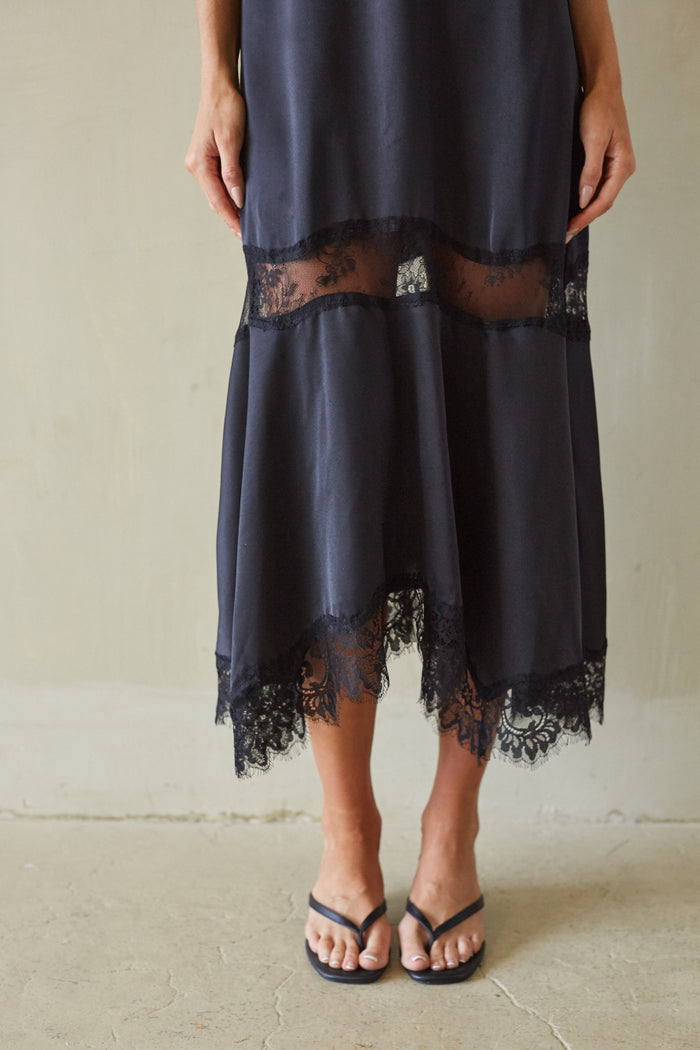 A person wearing a dark charcoal gray midi skirt with black lace trim and sheer mesh panels, paired with black flip-flop sandals, standing against a neutral beige wall.