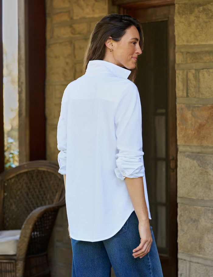 A woman wearing a white long-sleeve button-up shirt and blue jeans, standing on a stone porch next to a wicker chair.