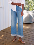 A person wearing light blue wide-leg cropped denim jeans with a rolled hem, styled with a white button-up shirt and tan flat sandals on a wooden deck.