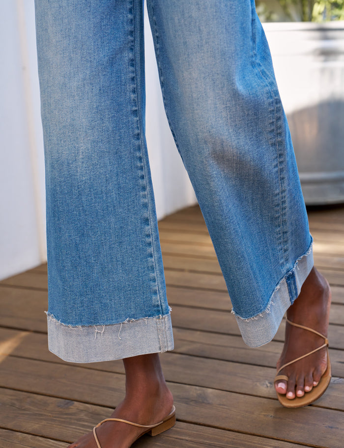 Close-up of a person wearing light blue wide-leg cuffed jeans with a raw frayed hem, paired with tan flat slide sandals on a wooden deck.