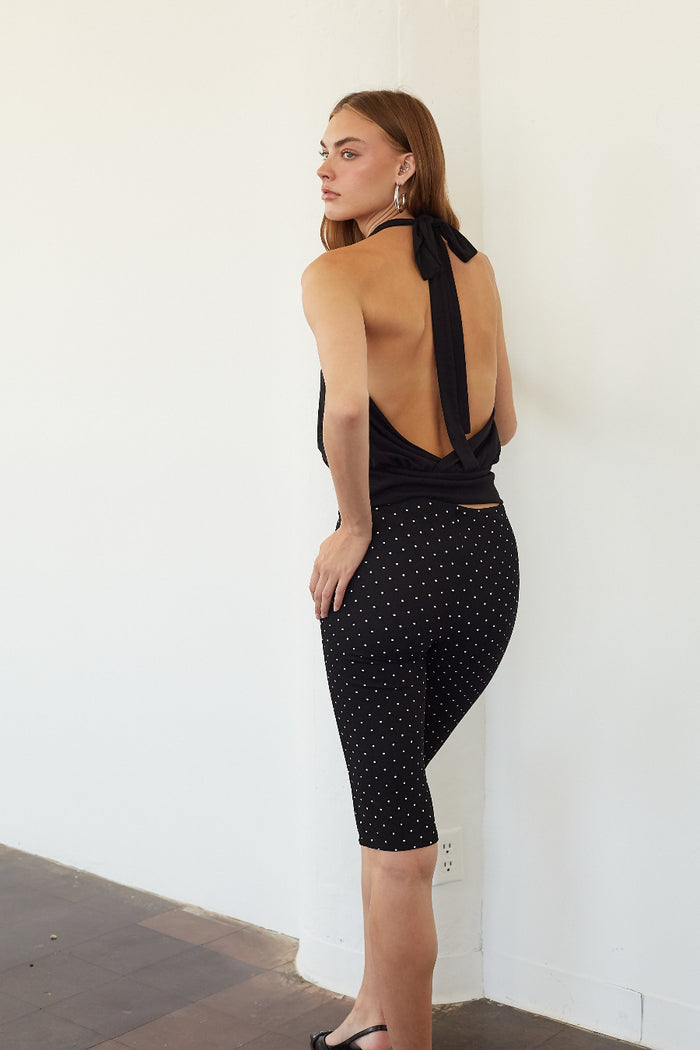 A woman wearing a black halter-neck top with a tie-back detail and matching black capri leggings with a small white polka dot pattern, standing against a white wall.