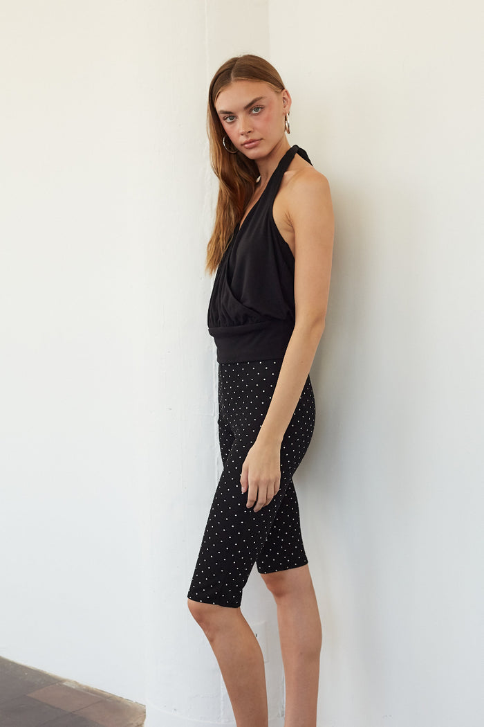A woman modeling a black halter top and black biker shorts with small white polka dots, standing against a plain white wall.