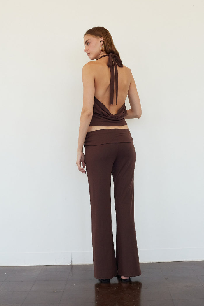A woman stands with her back to the camera wearing a dark brown two-piece outfit consisting of a halter-style wrap top and matching wide-leg pants, set against a plain white wall.