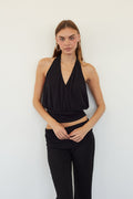 A woman modeling a black halter wrap-style crop top and black pants against a plain white wall.
