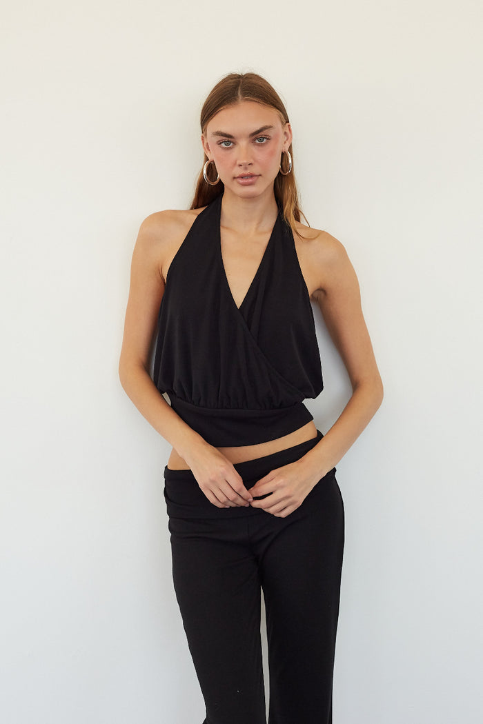 A woman modeling a black halter wrap-style crop top and black pants against a plain white wall.