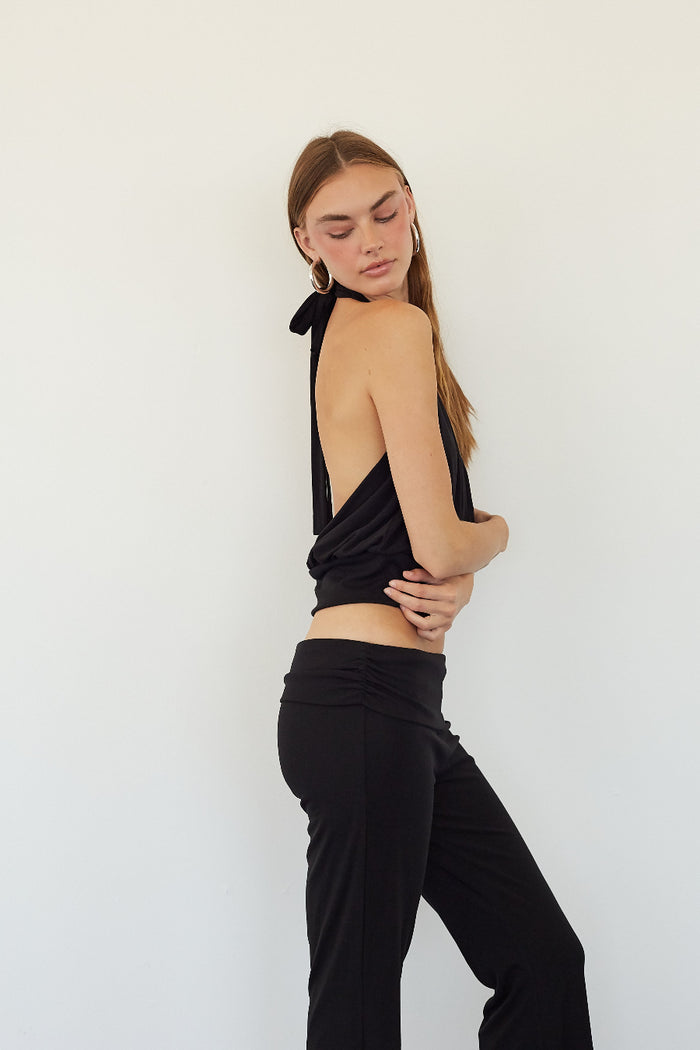A woman modeling a black halter crop top with a draped back and black pants, standing against a plain white wall.