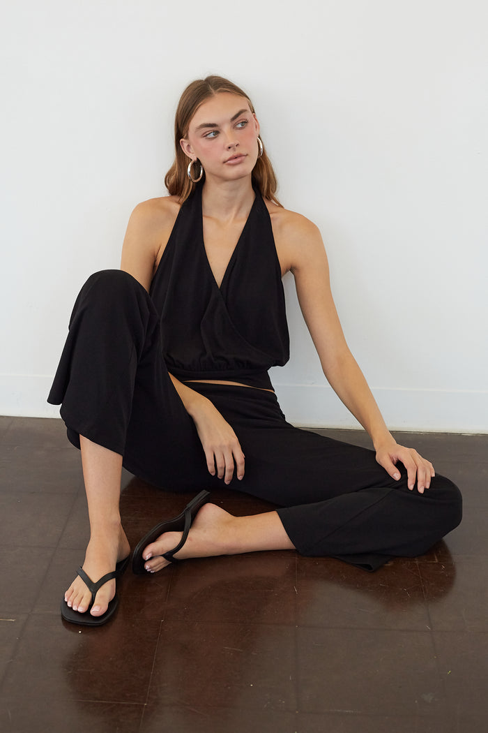 A woman sits cross-legged on a dark brown wooden floor against a plain white wall, wearing a black halter-neck wrap top and matching black cropped pants with black flip-flop sandals.