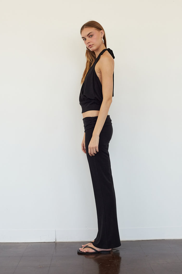 A woman stands in profile wearing a black sleeveless halter crop top and matching black wide-leg pants against a plain white wall.