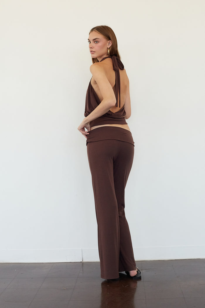 A woman stands in profile wearing a matching brown two-piece outfit consisting of a halter-style wrap top and high-waisted wide-leg pants, set against a plain white wall and dark wood floor.