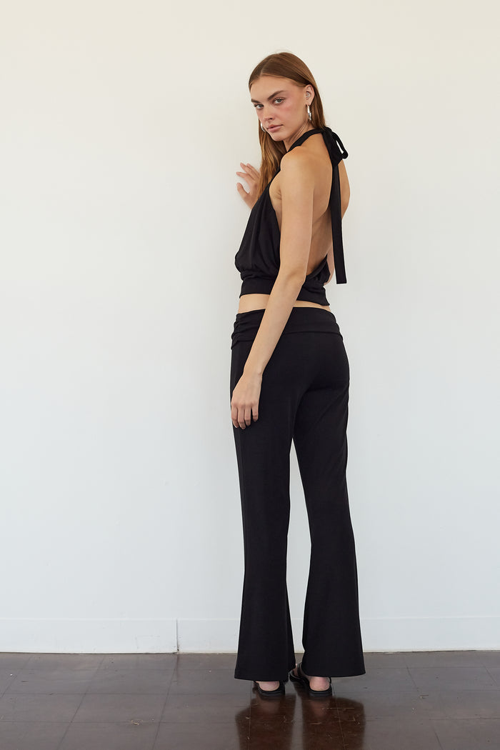 A woman stands in profile wearing a black halter crop top with a tie-back detail and matching black high-waisted flare pants, photographed against a plain white wall with a dark wood floor.