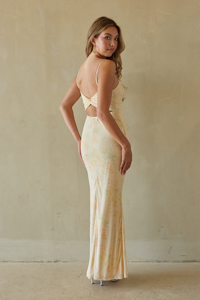 Woman wearing a long, light-colored dress with cut-out design against a plain wall.