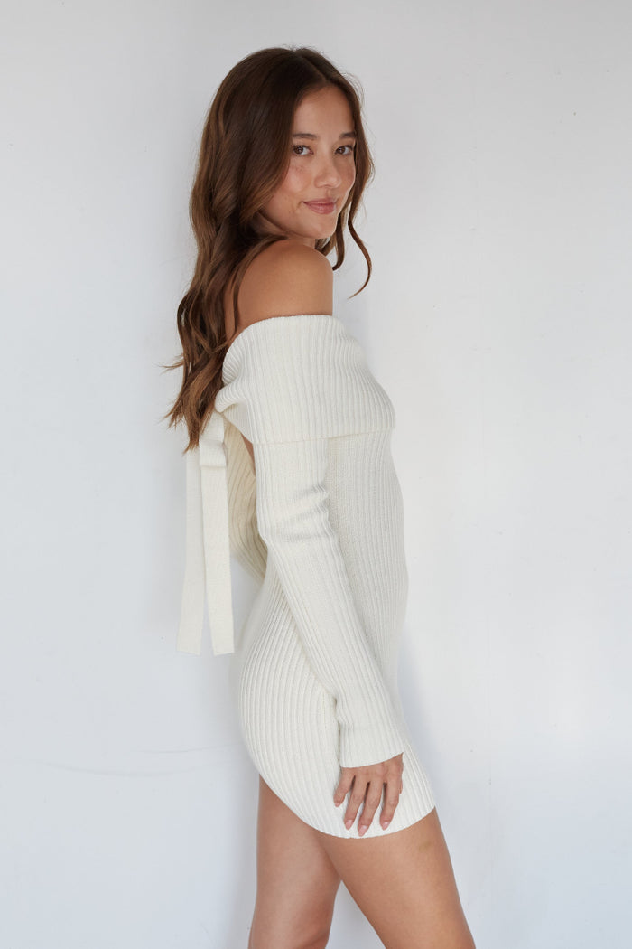 Woman wearing a white off-shoulder sweater dress against a plain background